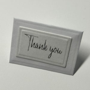 Puede incluir: Una tarjeta blanca con un borde en relieve y el texto "Gracias" escrito en cursiva negra.