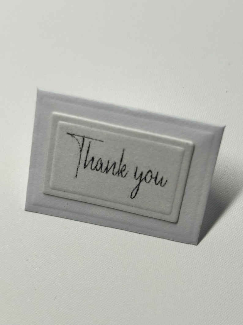 Puede incluir: Una tarjeta blanca con un borde en relieve y el texto "Gracias" escrito en cursiva negra.