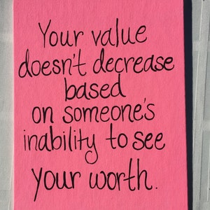 Puede incluir: Un letrero de lona rosa con letras negras que dice: "Tu valor no disminuye en función de la incapacidad de alguien para ver tu valía."