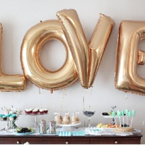 May include: Gold foil balloons spelling out the word "LOVE" are hanging from the ceiling above a table with a dessert spread. The table is decorated with teal and white accents.