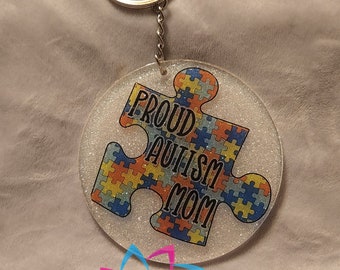 May include: A white round keychain with a glitter finish features a blue, yellow, and orange puzzle piece with the text "Proud Autism Mom".
