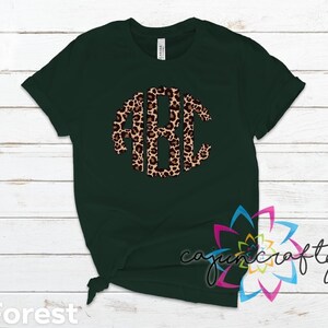 May include: A forest green t-shirt with a leopard print monogram in the center. The monogram is in the shape of a circle and features the letters A, B, and C.