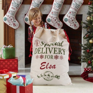 May include: A white canvas Santa sack with red and green trim. The sack has the text "Special Delivery for Elsa" printed on it. The sack is sitting on a floor in front of a fireplace with stockings hanging on it.