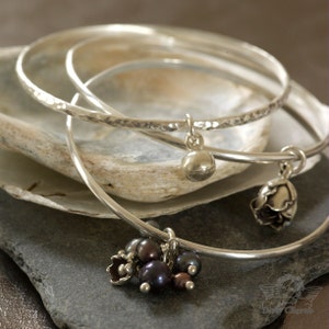 May include: Three silver bangles with hammered textures and dangling charms. The charms include a small ball, a flower with black pearls, and a stylized flower.