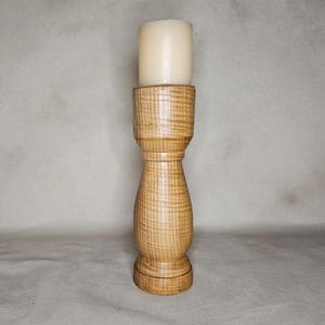 Op de afbeelding: Een hoge, cilindrische houten kandelaar met een lichtbruine, natuurlijke houtnerfafwerking en een gladde, gedraaide vorm. Een dikke, crèmekleurige stompkaars staat stevig in de bovenste uitsparing van de houten standaard.