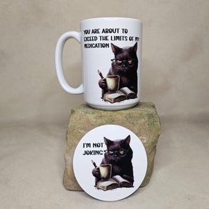 May include: A white ceramic mug and a round coaster featuring a black cat wearing glasses, holding a pencil and a cup, sitting by an open book. The mug text reads: YOU ARE ABOUT TO EXCEED THE LIMITS OF MY MEDICATION. The coaster text reads: I'M NOT JOKING.