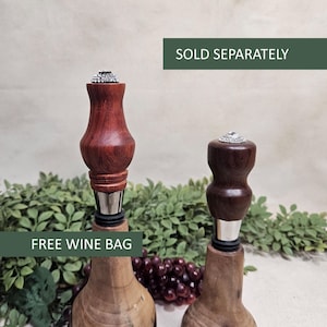 May include: Two wooden bottle stoppers with smooth, turned wood handles in reddish-brown and dark brown tones. Each stopper features a silver-colored metal base with black rubber rings and a small, circular, crystal-encrusted accent on the top.
