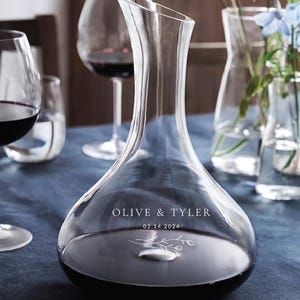 Puede incluir: Una decantador de vidrio transparente con una base ancha y un cuello estrecho. El decantador está lleno de vino tinto y tiene el texto "Olive & Tyler 02.14.2024" grabado en el lateral.