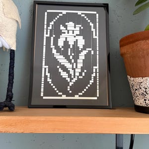 May include: A rectangular black frame containing a piece of art with a white pixelated floral design on a black background. The artwork features a stylized flower centered within an oval border, all composed of small square white segments.