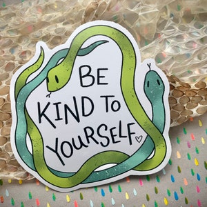 Könnte beinhalten: Ein Aufkleber mit zwei grünen Schlangen, die sich ineinander verschlungen haben und eine Herzform bilden. Der Text "Be Kind to Yourself" ist in Schwarz innerhalb des Herzens geschrieben.
