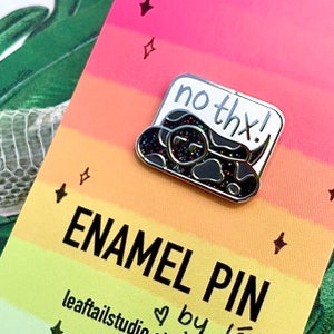 Könnte beinhalten: Eine schwarze und silberne Emaille-Anstecknadel mit einer Cartoon-Schlange mit einer Sprechblase, auf der "no thx!" steht. Die Anstecknadel befindet sich auf einem Regenbogenhintergrund mit Sternen.
