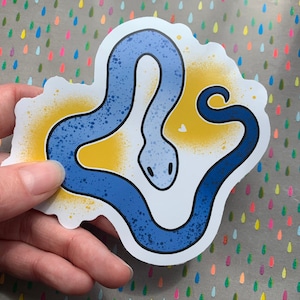 Könnte beinhalten: Ein blauer und gelber Aufkleber mit einem Cartoon-Schlangen-Design. Die Schlange ist zu einem Kreis zusammengerollt, mit dem Schwanz im Maul.
