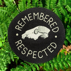 May include: A round, black patch with the words "REMEMBERED RESPECTED" in white, encircling a white illustration of an animal skull. The patch is set against a backdrop of green fern leaves.