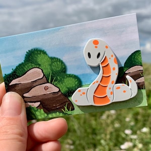 May include: White enamel pin of a cartoon snake with orange spots. The snake is curled up with its head facing the viewer. The pin is on a card with a cartoon background of a green bush and brown rocks.