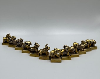 May include: Twelve gold-coloured animal figurines, each representing a different Chinese zodiac sign, are arranged in a semi-circle on a white surface. The figurines are detailed and have a traditional Chinese style.