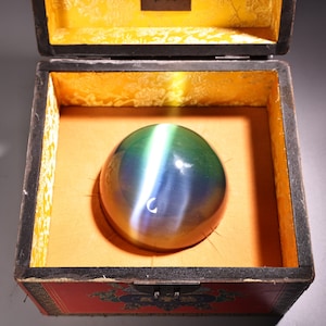 Op de afbeelding: Een grote, iriserende, groene en blauwe edelsteenbol wordt getoond in een houten doos met een rode en gouden voering. De bol ligt op een zacht, oranje kussen.