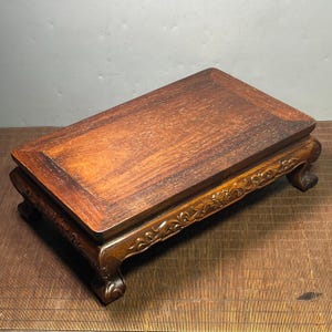 May include: A rectangular, dark brown wooden display stand with a raised, framed top. The stand has carved floral designs along the apron and curved legs. The wood grain is visible on the surface.