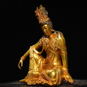 May include: A golden statue of a seated figure with ornate details, including a crown and jewelry. The figure is in a relaxed pose, with one arm extended. The statue has a polished finish and is set against a dark background.