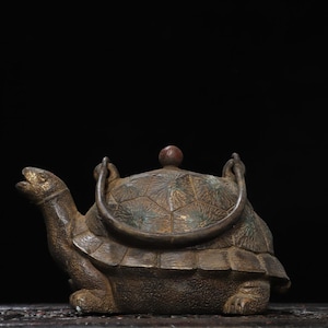May include: A bronze-toned metal teapot shaped like a turtle with a textured shell pattern, a curved handle attached to the top, and a small spherical knob on the lid. The turtle head is extended forward with an open mouth, and the body has a rough finish.