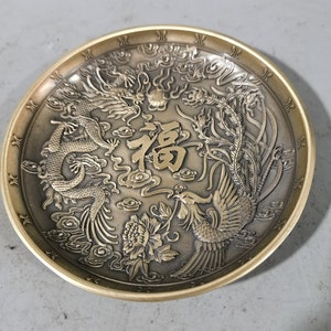 Op de afbeelding: Een ronde, messingkleurige plaat met een ingewikkeld ontwerp met een draak en een feniks. De plaat is versierd met een bloemmotief en het Chinese karakter voor "geluk".