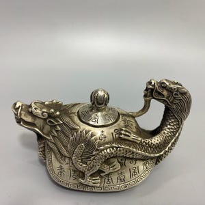 Puede incluir: Una tetera plateada con un diseño de dragón. La boquilla es una cabeza de dragón, y el cuerpo del dragón forma el asa. La tapa tiene un pequeño pomo y caracteres chinos grabados en el cuerpo.