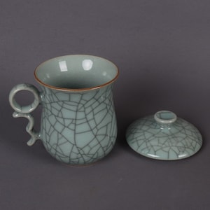 May include: A light blue ceramic teacup with a crackle glaze and a matching lid. The teacup has a handle in the shape of a loop.