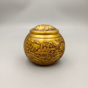 Op de afbeelding: Een gouden metalen container met een deksel, met een ingewikkeld reliëf van een traditioneel Chinees landschap.