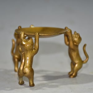 Könnte beinhalten: Ein dekorativer Ständer aus goldenem Metall mit einer runden Oberseite und drei Katzenbeinen. Die Katzen halten die Oberseite des Ständers hoch.
