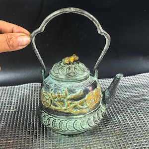 Puede incluir: Tetera antigua de bronce con asa curva. La tetera presenta un diseño detallado con una rana en la tapa y motivos de dragones y monedas. La tetera tiene un pico y mide aproximadamente 15 cm de altura.