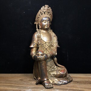 May include: A seated bronze statue with a serene expression, adorned with intricate details. The figure wears a decorative headdress and draped garments, with hands clasped around a sphere. The statue has a weathered, antique appearance.