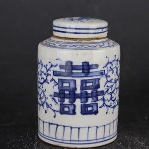 Könnte beinhalten: Eine weiß-blaue Keramikdose mit Deckel. Die Dose zeigt ein blaues Doppelglück-Symbol und florale Muster. Der Deckel hat blaue Streifen und ein florales Design. Die Dose ist zylindrisch mit leicht abgerundetem Boden.