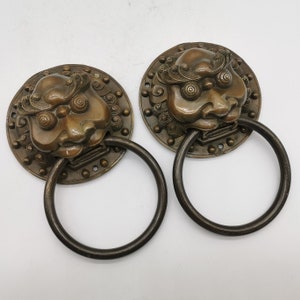 Puede incluir: Dos aldabas de puerta de bronce antiguo con diseños de cabeza de león. Cada aldaba tiene una base circular con detalles intrincados y un asa de anillo.