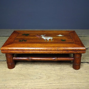 May include: A small, square wooden table with a rich, reddish-brown finish. The tabletop features inlaid decorative elements, including animal and plant motifs. The table has four short, rounded legs and a bamboo-style support beam, creating a unique aesthetic.