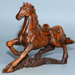 Op de afbeelding: Een handgesneden houten paardenbeeld met een zadel en ingewikkelde details. Het paard is afgebeeld in een galoppose met zijn hoofd naar de zijkant gedraaid. Het beeld is gemaakt van donkerbruin hout en heeft een natuurlijke afwerking.