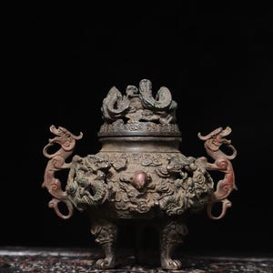 May include: A detailed bronze incense burner, featuring dragon and cloud motifs. The burner has two dragon handles and a lid adorned with more dragon figures. The color palette includes bronze and reddish-brown tones, set against a dark backdrop.