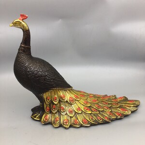 Op de afbeelding: Een bronzen pauwsculptuur met een gouden en rode staart. De pauw heeft een donkerbruin lichaam, een gouden kop en een rode kuif. De sculptuur is een decoratief item.