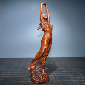 Op de afbeelding: Een houten sculptuur van een vrouw in een vloeiende jurk, met haar armen boven haar hoofd geheven. Het beeld is gesneden uit roodbruin hout en heeft een gedetailleerde, realistische stijl.