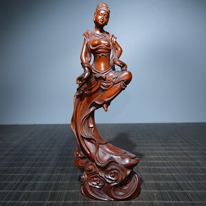Op de afbeelding: Een handgesneden houten beeld van een vrouw in een vloeiende jurk, staande op een wolkvormige basis. Het beeld is gemaakt van donker hout en heeft een gedetailleerd ontwerp.