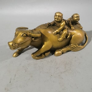 Op de afbeelding: Een gouden metalen sculptuur van een waterbuffel met twee kinderen die op zijn rug rijden. De buffel ligt met zijn hoofd naar de zijkant gedraaid. De kinderen lachen en hebben hun armen om de nek van de buffel geslagen.