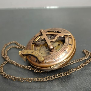 May include: A vintage brass sundial compass with a hinged lid and a chain. The compass features etched markings and a gnomon. The sundial is a warm, antique gold colour.