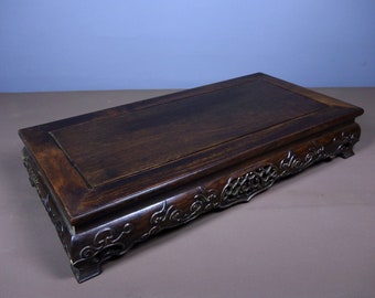 May include: A dark brown wooden display stand with intricate carvings on the base and a smooth, rectangular top.