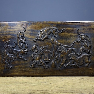 Op de afbeelding: Een rechthoekig houten snijwerk met een gedetailleerd reliëf van tijgers en een figuur. De tijgers worden in verschillende poses afgebeeld en de figuur houdt een voorwerp vast. Het hout heeft een donkere, verouderde uitstraling.