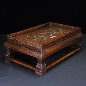Può includere: Un espositore rettangolare in legno con finitura marrone scuro. L'espositore presenta un motivo floreale intagliato lungo i bordi e un intarsio decorativo sulla superficie superiore. È sostenuto da quattro gambe curve.