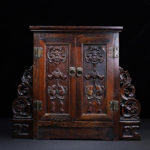Op de afbeelding: Een donkerbruine houten kast met twee deuren en sierlijke houtsnijwerken. De kast heeft messing scharnieren en ringtrekkers. Decoratieve gebeeldhouwde panelen flankeren de kast en dragen bij aan de antieke uitstraling.