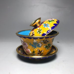 May include: A small, yellow teacup with a lid, decorated with colourful flowers and butterflies. The teacup and lid are made of cloisonné, a traditional Chinese enamelware technique.