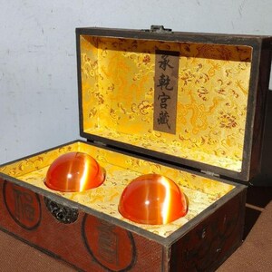 Puede incluir: Dos esferas de vidrio naranja en una caja de madera con un forro estampado en oro y negro. La caja tiene un cierre negro y el texto "安 鎮 吾 願" está escrito en una etiqueta de papel dentro de la caja.