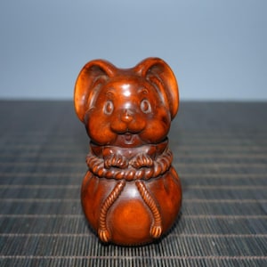 Op de afbeelding: Een houten beeldje van een muis die een zak vasthoudt. De muis is bruin en heeft een glimlachend gezicht. De zak is vastgebonden met een touw.