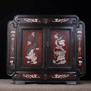 May include: A dark wood cabinet with two doors and a drawer. The doors feature red panels with inlaid mother-of-pearl dragons and phoenixes. The cabinet has decorative carvings and metal hardware.
