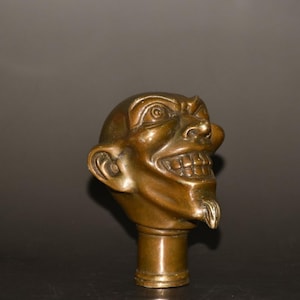 May include: Bronze-colored cane handle shaped like a grinning face with exaggerated features. The face has a large nose, wide grin, and a small goatee. The handle is cylindrical and appears to be made of metal.