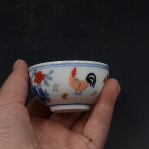 May include: Small white ceramic bowl with a hand-painted rooster and floral design. The rooster is red, orange, and yellow with a black tail. The bowl has a blue rim and is held in a hand.